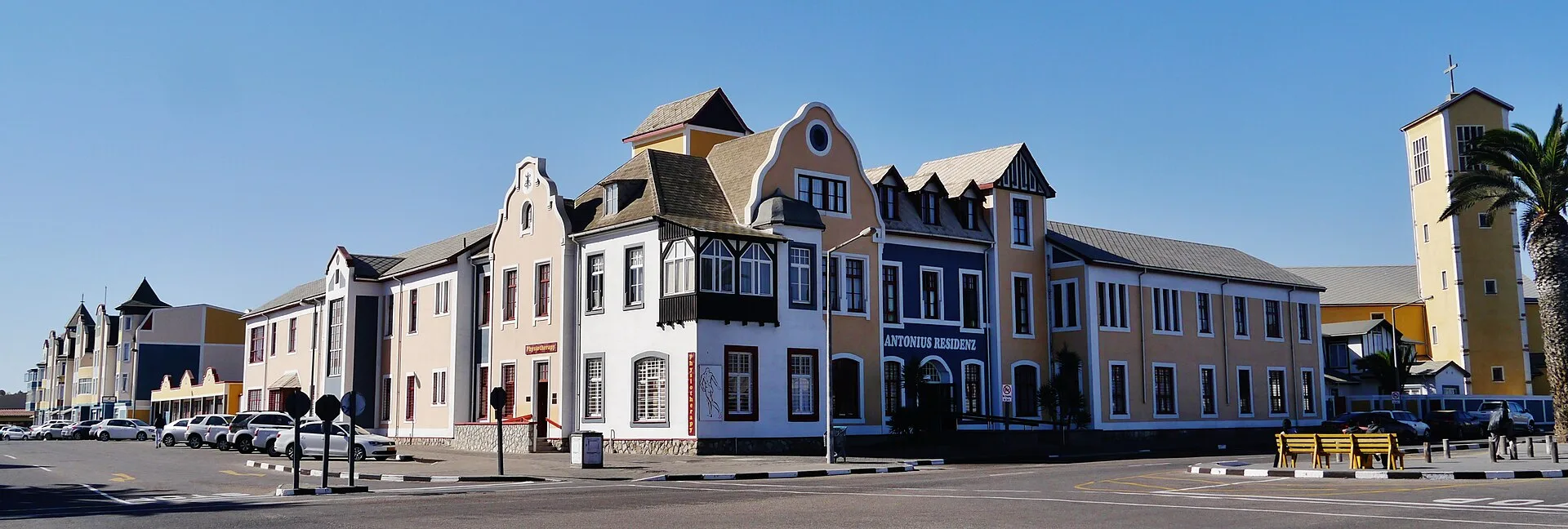 Rue d'un quartier avec des façades de style colonial allemand colorées, une résidence et un clocher jaune sous un ciel bleu